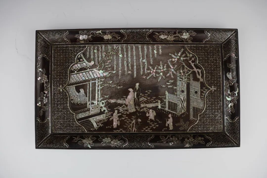 清黑漆嵌螺钿庭院人物图长方盘 通体髹黑漆为地,嵌螺钿为饰。盘心钱纹锦地开光内,饰有庭院人物图。只见亭台竦峙,俢竹玉立,柳丝摇曳,翠鸟飞舞;文人雅士汇聚一堂,或赏景赋诗,或谈玄论道,其旁有童子携琴而立,文心雅趣之味甚浓。黑漆之幽玄深谧与螺钿之五彩绚丽巧妙结合,构成了螺钿漆器独有的美感与趣致。
