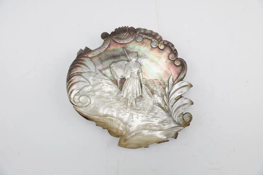 20世纪贝雕“赶海女”图摆件 通体采用雕刻工艺,中心以减地浮雕的技法雕琢出西洋赶海女的形象。只见其身背竹篓,手持抄网,站立于海滩之上眺望远方。贝雕上方浮雕如意海水纹,左右两侧饰海草,下方饰鱼纹。贝雕摆件往往因材施艺,既保有贝壳的天然质感,又展现了精湛的雕工。